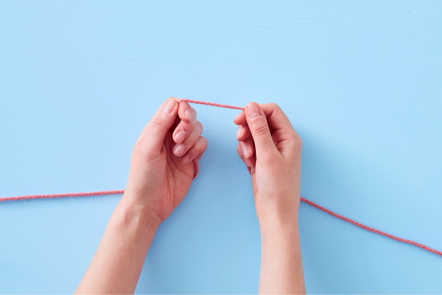 A series of images demonstrating how to create your first stitch. They show an overhead view of two hands using a grey metal knitting needle and pink yarn, on a blue background.