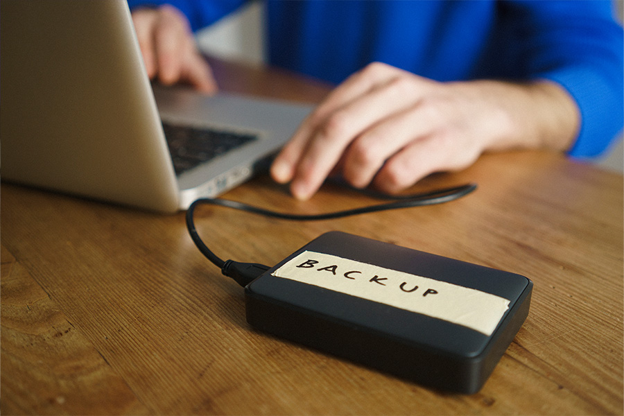 A person sitting at a desk using a laptop with a portable data storage device labelled ‘backup’ connected.