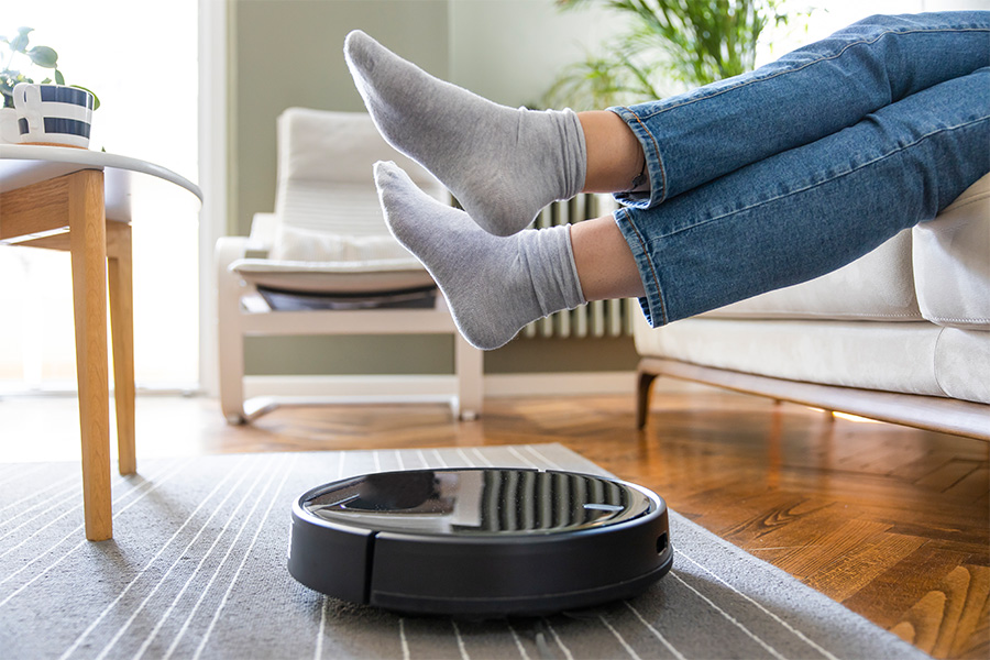 A robot vacuum cleaner operating on a rug and wooden floor with a person sitting on a lounge nearby in a living room setting.