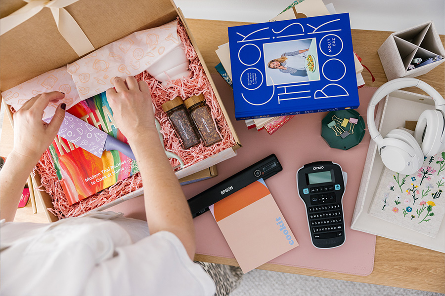 An overhead shot of Emily arranging items in a gift box at a desk, with cookbooks, a label maker, a portable scanner and headphones also on the desk.
