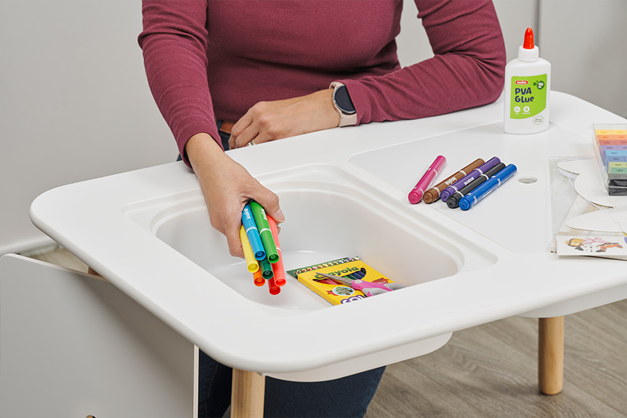 Pip Renfrew sitting at a kids’ craft desk puts away craft items including Crayola marks, PVA glue and scissors to declutter the table. 