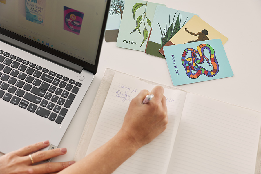 An open laptop sits on a desk next to a notebook with hands writing on the blank pages. Above the notebook are some Riley Callie Resources flash cards.