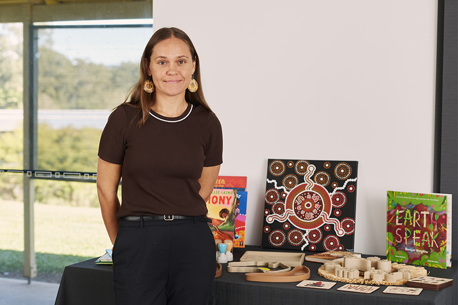 A woman wearing a black t-shirt and pants stands in front of a white wall and some windows. Behind her is a table with a black tablecloth, with books, Indigenous artworks and wooden stamps on top of it.