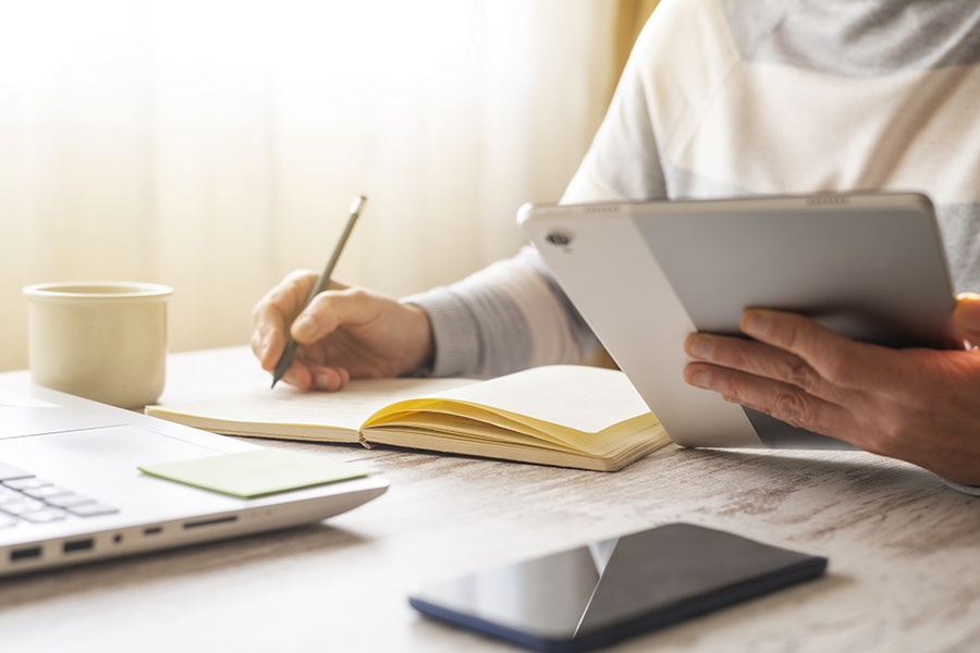 A person sitting at a desk with a laptop, mug and smartphone in front of them, and holding a tablet while writing in a notebook.