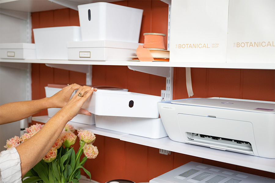 Ange Norman organising storage boxes on a shelf, with a printer and decorative items nearby.