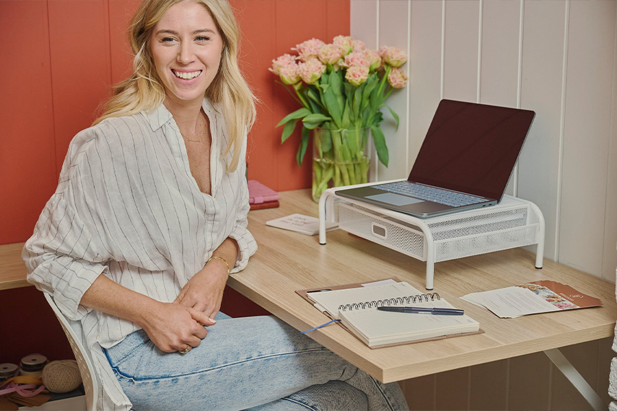 Ange Norman sitting at a wooden desk with a laptop on a stand, a notebook and a vase of pink flowers.