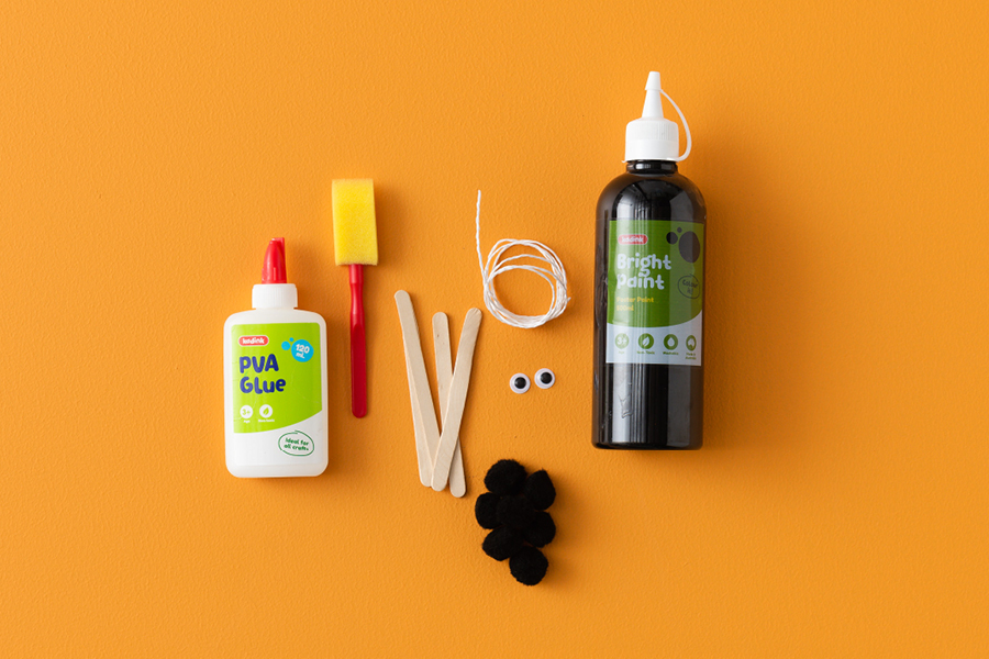 An overhead view of products that can be used to make a spooky spider, including PVA glue, a paint sponge, craft sticks, black paint, black pom-poms, string and googly eyes.