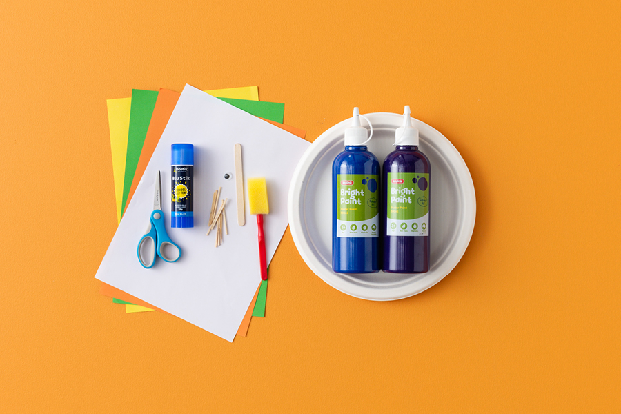 An overhead view of products that can be used to make a flying witch plate, including a stack of coloured paper, scissors, glue, matchsticks, a disposable plate, paint and a paint sponge.
