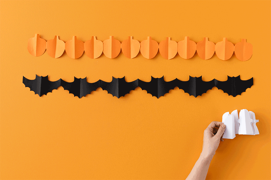 A series of images showing how to make paper garlands in the shape of a pumpkin, a bat and a ghost for a Halloween craft project for kids.