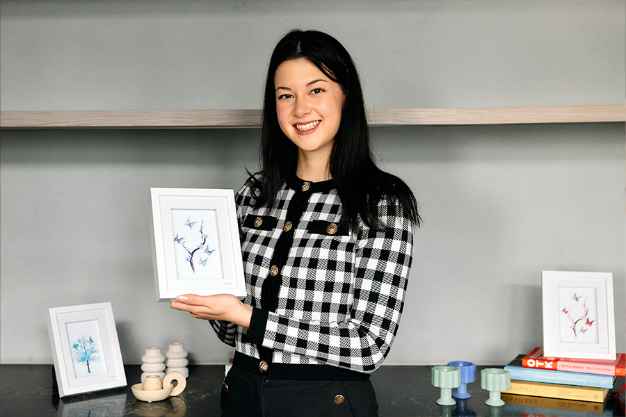 Kiana Mei holding a framed artwork featuring origami cranes, with additional framed art, books, and decorative items displayed on shelves in the background.