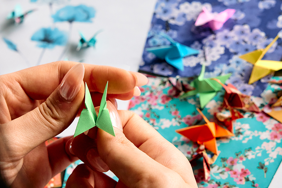 A close-up of hands folding a green origami crane, surrounded by colourful paper cranes and floral-patterned paper on a crafting table.