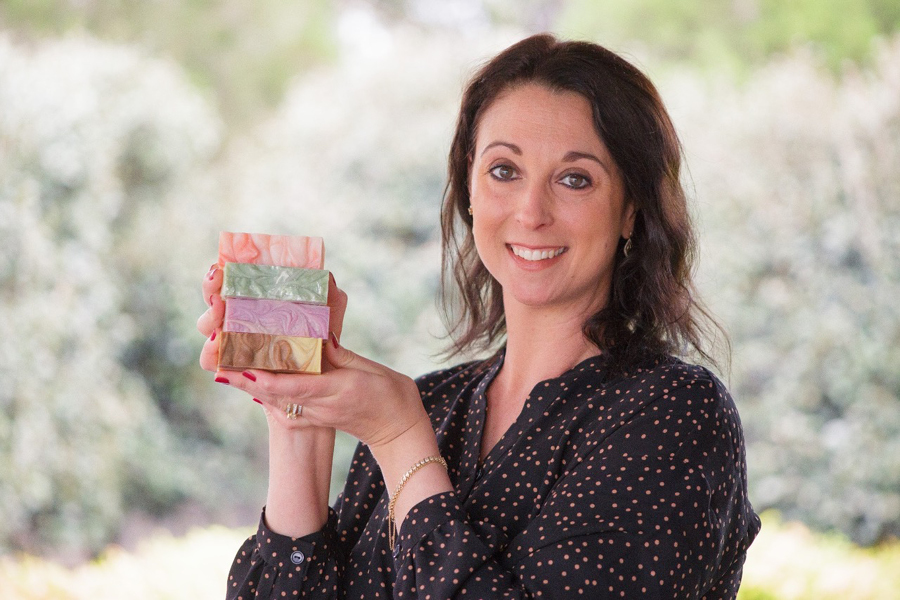 L’ada Soaps & Scents founder Deborah Terkely holding a stack of four soaps in various colours. She’s standing outside with leafy green bushes out of focus in the background.