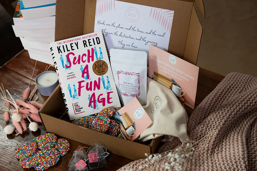 A cardboard box filled with tea, chocolates, a book, calico bag and printed cards and vouchers sits on a wooden table surrounded by a blanket, a pile of books, a candle and flowers.