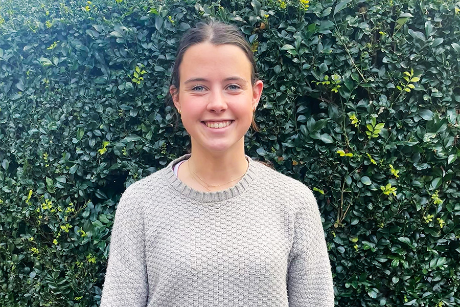 A close-up shot of Lucy Molineux standing outdoors in front of a lush green hedge.