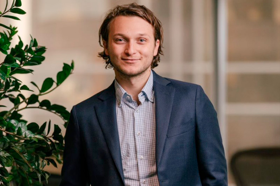 A close-up shot of Max Milstein standing indoors next to a green plant with a modern office background.