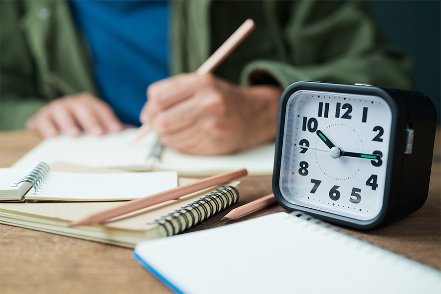 A close-up of a desk with a clock, notebooks and pencils, while a person writes in the background.