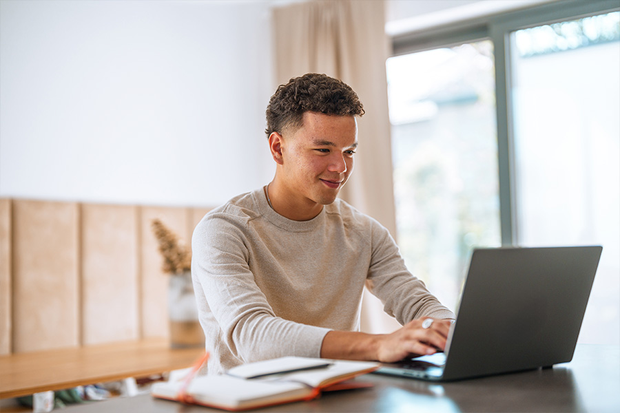 A student using a laptop at a desk in a bright, modern room, with a notebook and pen nearby.