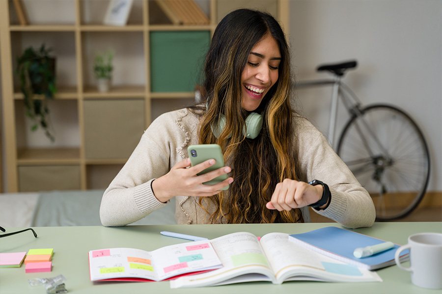 A student sitting at a desk with a smartphone, an open notebook and textbook, sticky notes, in a room with shelves and a bicycle in the background.