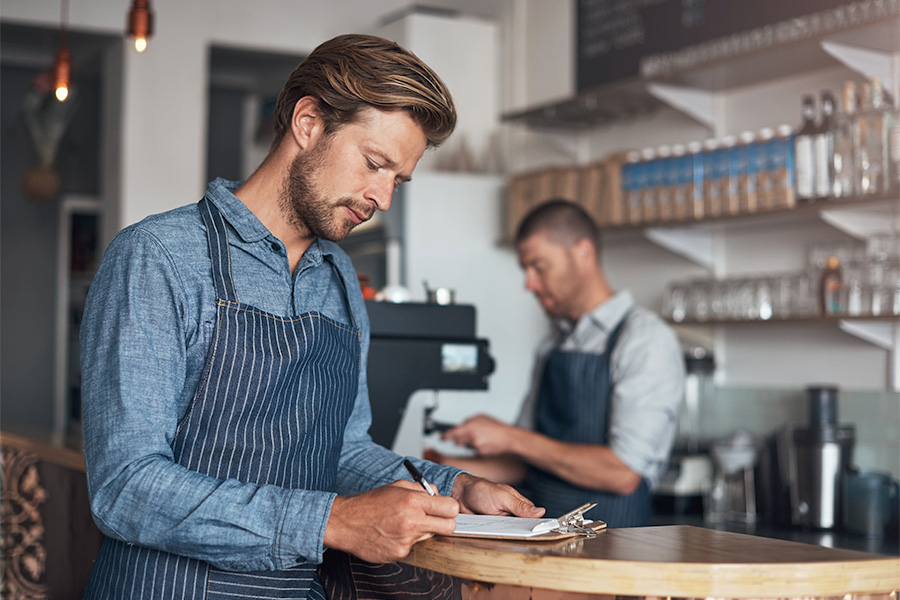 A person wearing a striped apron writing on a clipboard at a cafe counter, with another person working at a coffee machine in the background.