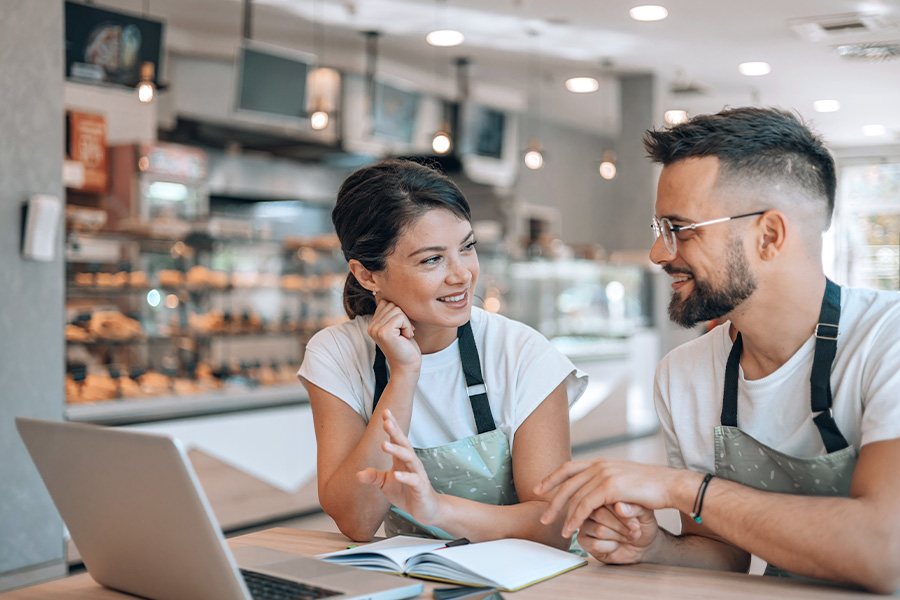 Two people in aprons sitting side by side and talking at a table with a laptop and notebook, in a cafe setting.
