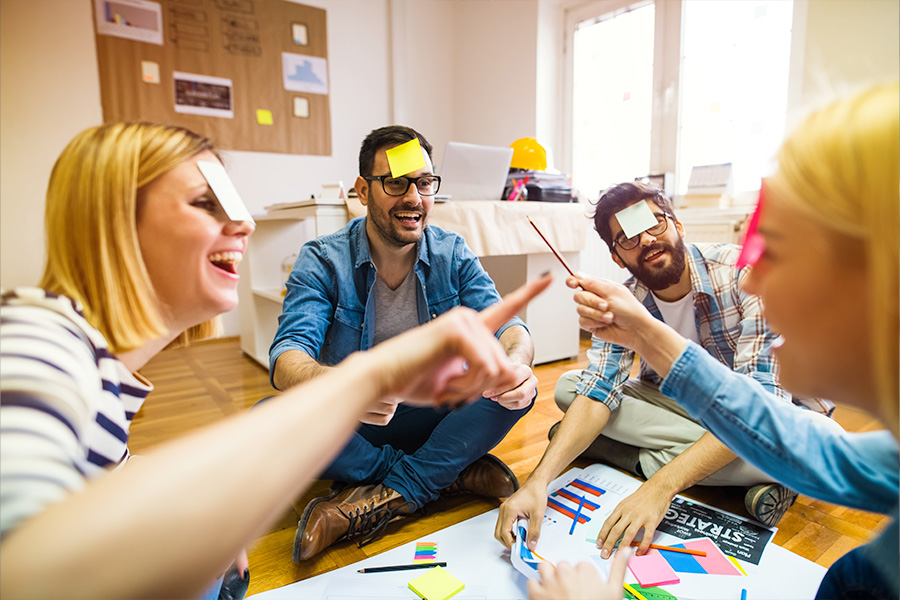 A group of office workers participating in a team activity. They are sitting on the floor in a circle with sticky notes stuck to their foreheads.
