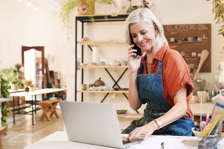 A small business owner working on a laptop and talking on the phone in a bright, creative studio filled with pottery and artistic supplies.
