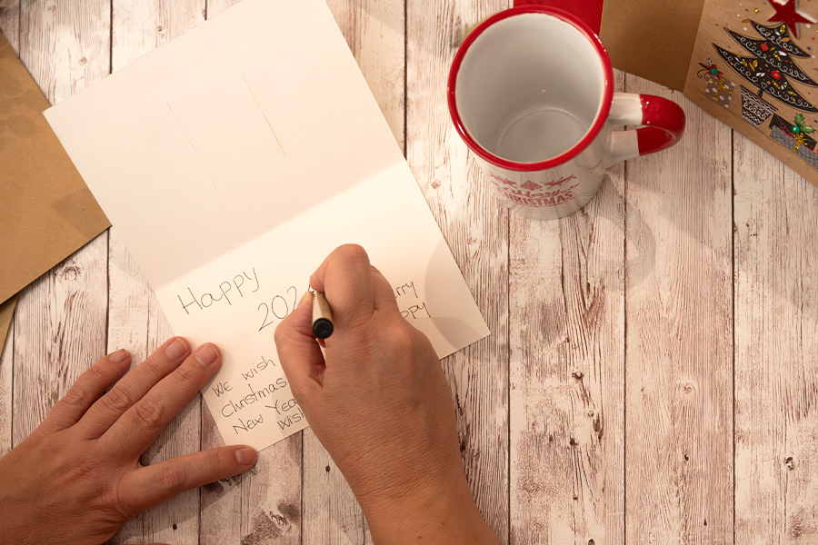 Person writing in a holiday card on a wooden table with a festive mug and Christmas decorations nearby.