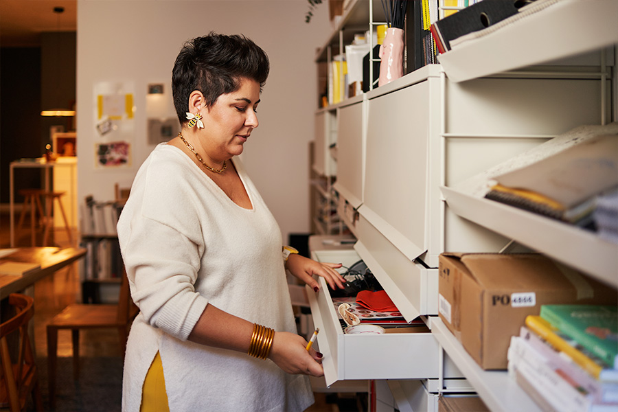 A person organising items in a drawer within a modern, well-lit workspace featuring desks and shelves filled with neatly organised books, folders and stationery.