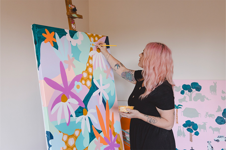 Rach Jackson painting a large, colourful floral artwork on a canvas in a studio, holding a paintbrush and palette, with vibrant designs visible in the background.