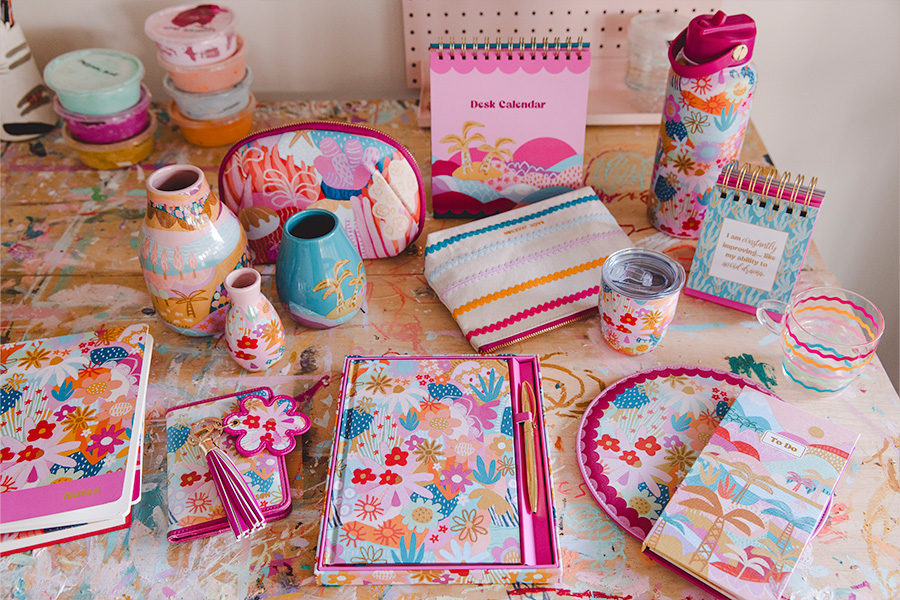 Items from the Officeworks Rach Jackson collection, including stationery, ceramics and accessories featuring vibrant floral and tropical designs, displayed on a paint-splattered table in an artistic setting.