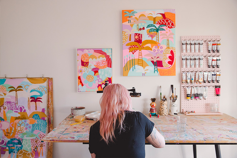 Rach Jackson working at a paint-splattered table in a studio, surrounded by colourful paintings, art supplies and tools organised on a pegboard and shelves.