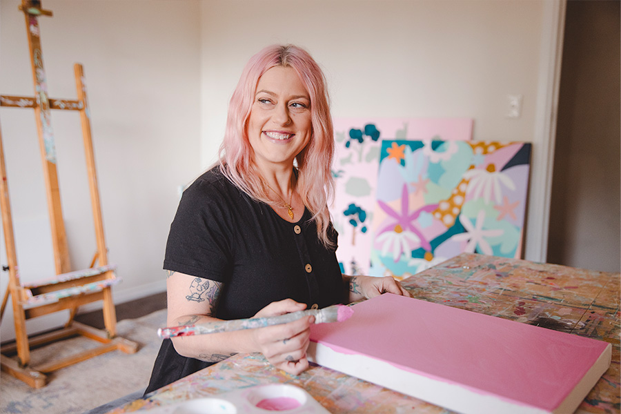 Rach Jackson painting a canvas with pink paint at a table in a studio, surrounded by vibrant artworks and an easel in the background.