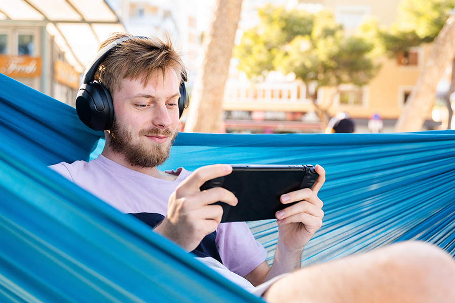 A person relaxing in a blue hammock outdoors, wearing headphones and playing on a handheld gaming device, with trees and buildings visible in the background.