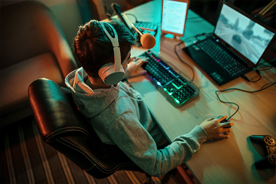 A person wearing headphones sitting in a gaming chair, using a keyboard and mouse at a desk with a gaming laptop and monitor displaying a racing game.