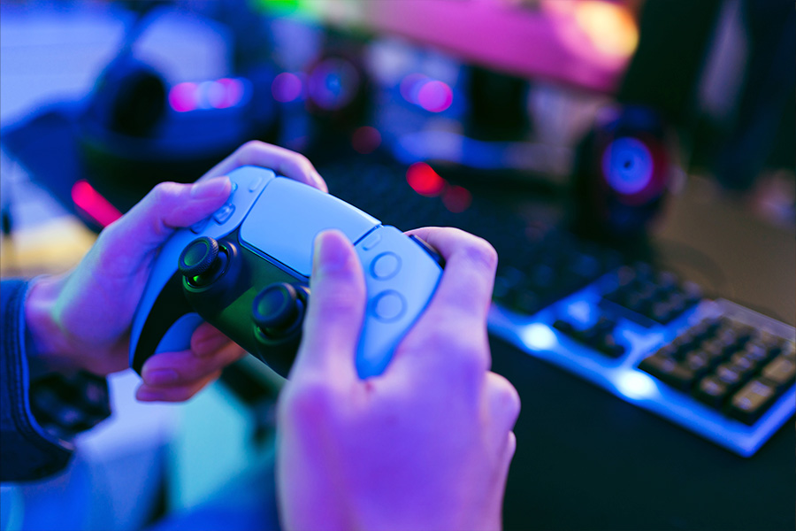 A close-up of hands holding a gaming controller in a brightly lit setup with a keyboard, headphones and other gaming equipment blurred in the background.