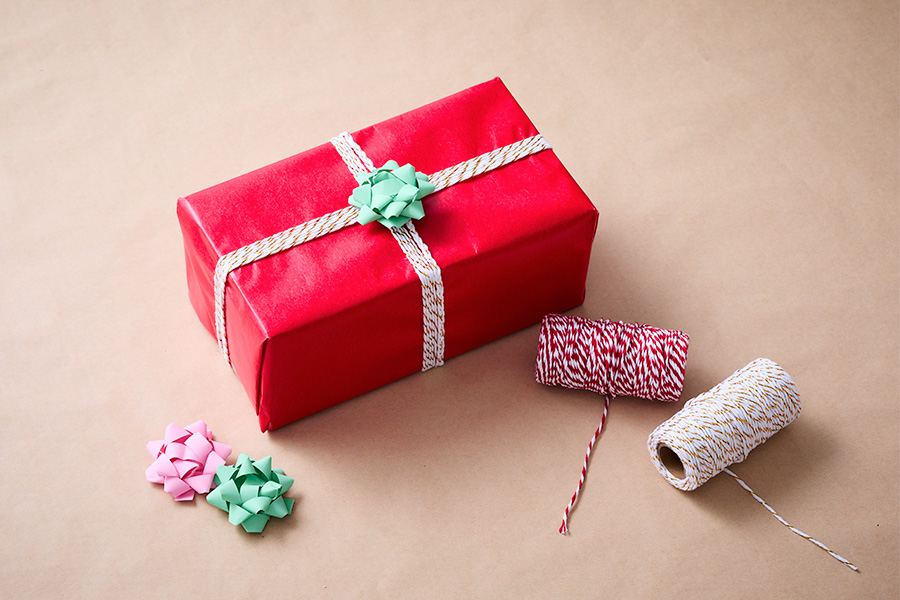 A present neatly wrapped in red gift wrapping, tied with orange and white twine and adorned with a green bow, surrounded by rolls of twine and additional pink and green bows on a plain brown background.