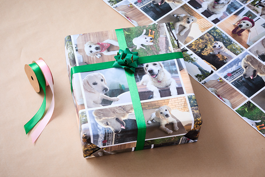 A gift wrapped in custom wrapping paper featuring photos of a golden retriever, tied with a green ribbon and bow, placed alongside rolls of pink and green ribbon on a brown background.