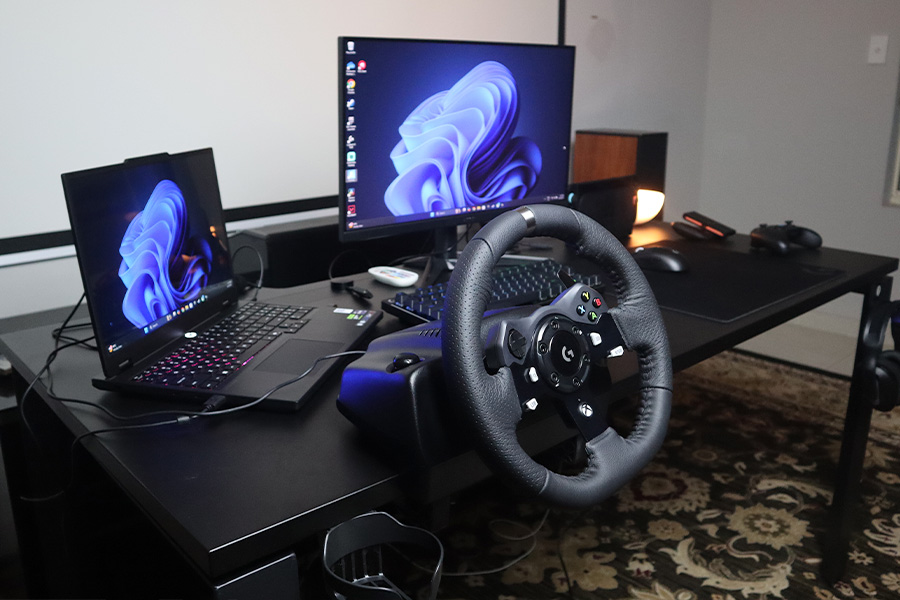 A gaming setup featuring a black sit-stand gaming desk, a racing wheel, a laptop and a monitor, keyboard and mouse, a wireless controller and lamp.