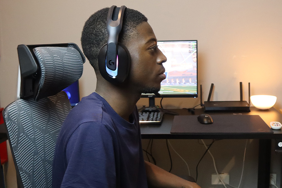 A close-up, side-on shot of Izzy Ngoy wearing a gaming headset, a gaming setup, including a monitor displaying a game, a keyboard, mouse, wi-fi router and a glowing lamp on a desk is visible in the background.