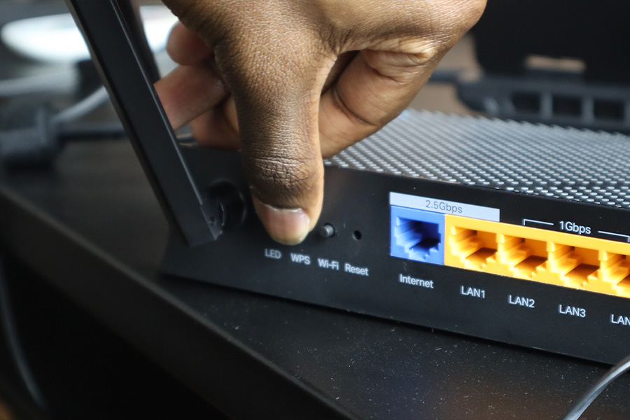A close-up of a hand pressing the WPS button on a wireless router, showing labelled ports for Internet, LAN connections and a 2.5Gbps port on a black desk.