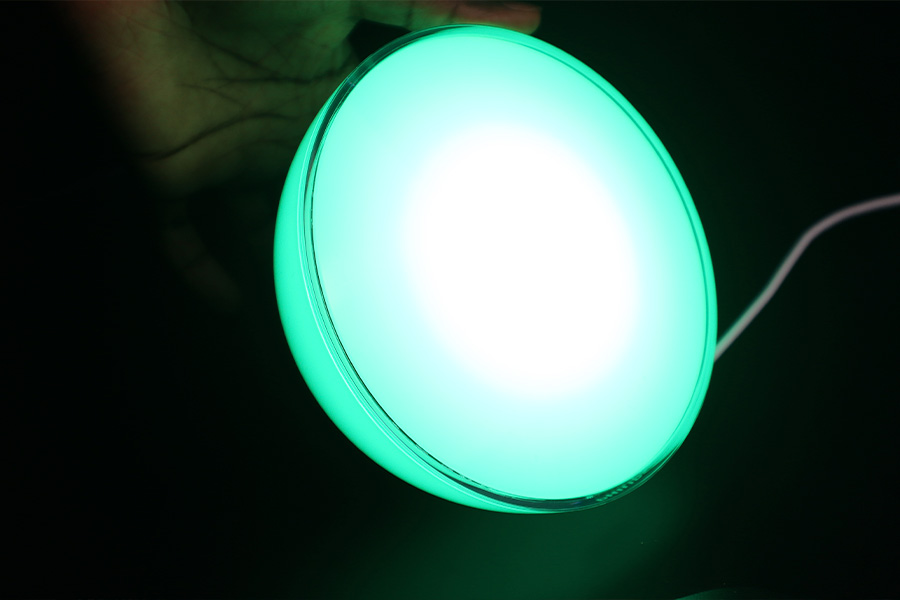 A glowing circular lamp emitting a bright green light, held in a hand against a dark background, with a white power cord visible.