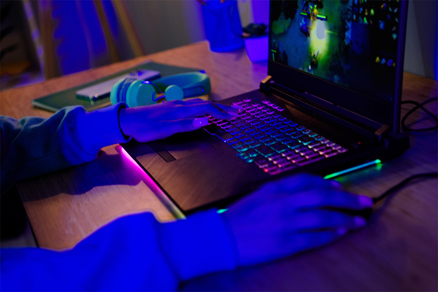 A close-up of hands on a rainbow backlit keyboard of a gaming laptop and mouse, playing a video game. A phone and a gaming headset sit on the desk nearby.