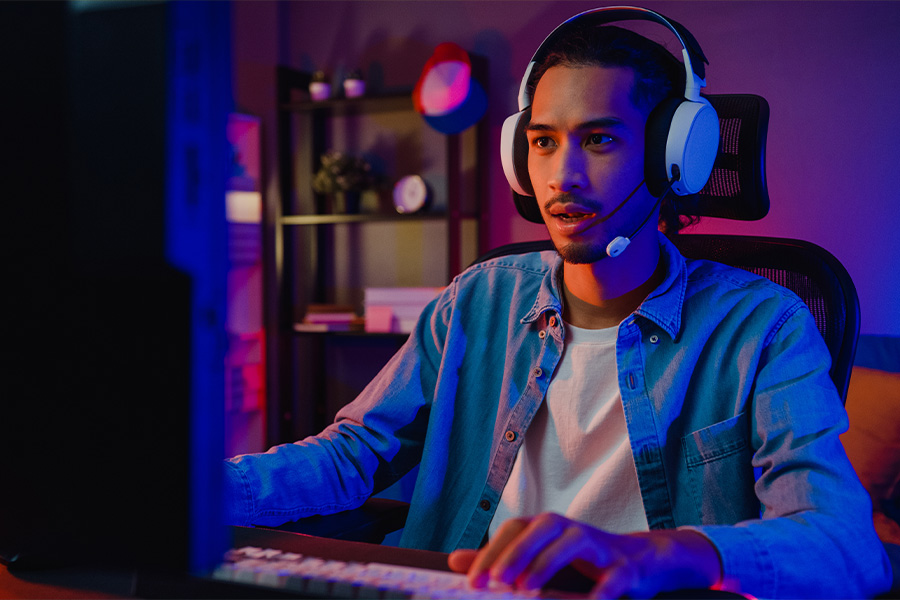 A man wearing a gaming headset intensely focused on playing a game on a large desktop display. The room is lit with colorful lights, creating a cool gaming atmosphere.