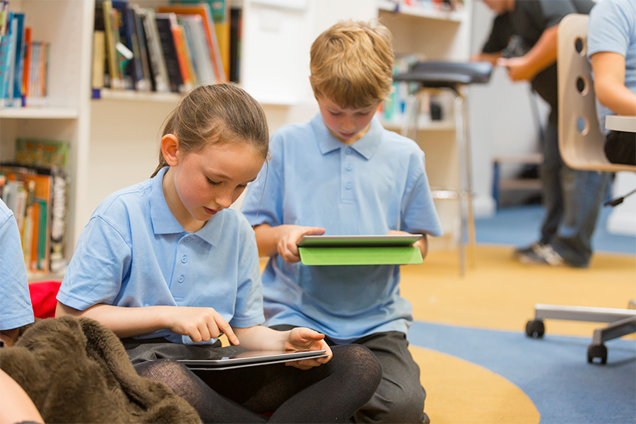 Two primary school students sitting on the floor of a classroom, engaging with tablets, surrounded by bookshelves and educational materials.