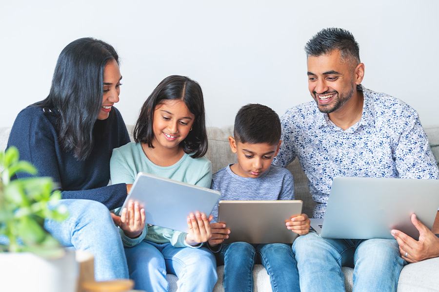 Two adults sitting on a couch with two children, engaging with digital devices including tablets and a laptop, with a plant visible in the foreground.