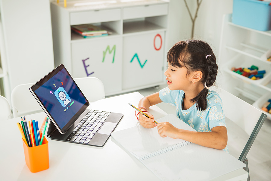 A primary-school-aged child sitting at a desk with a tablet displaying an educational app, holding a pencil and writing in a notebook, surrounded by colourful stationery in a bright classroom.