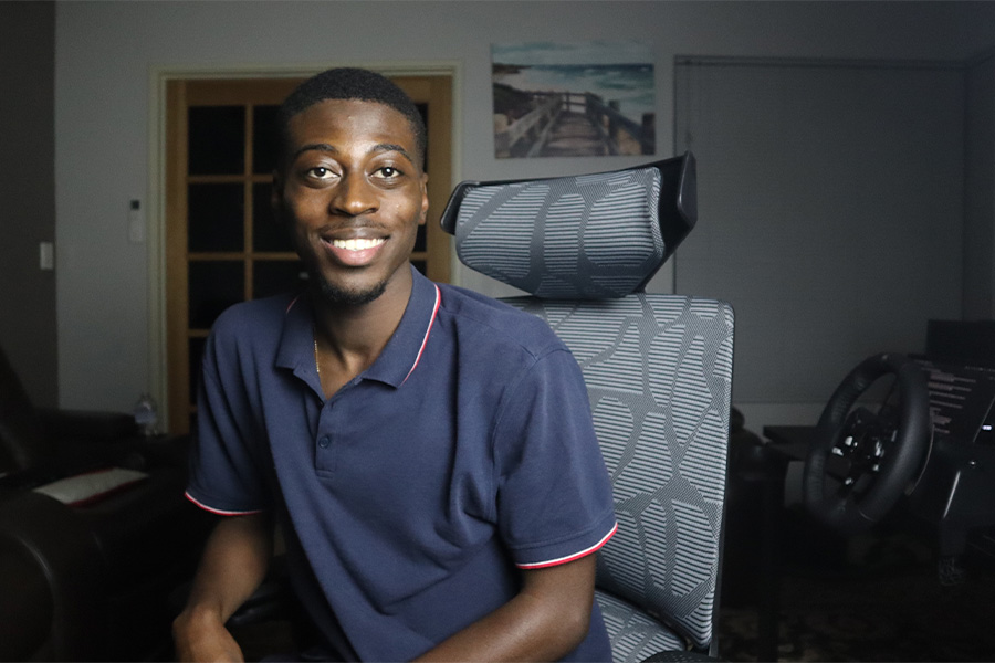 Izzy Ngoy sitting in an ergonomic, patterned office chair in a dimly lit room, with gaming equipment including a racing wheel setup visible in the background.