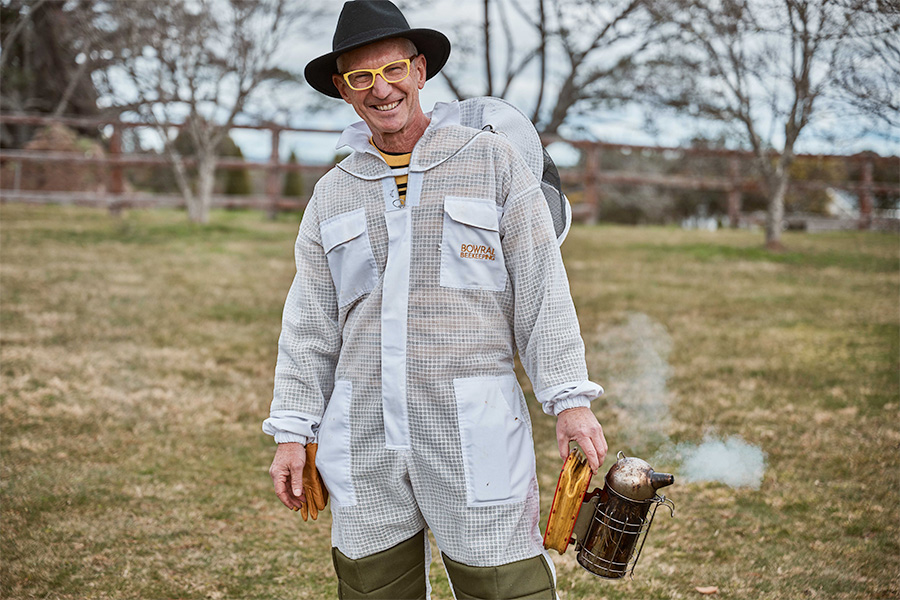 Founder of Bowral Honey Farm, Hamish Ta-mé, wearing a beekeeper suit and holding a smoker in a grassy outdoor setting with trees and fencing in the background.