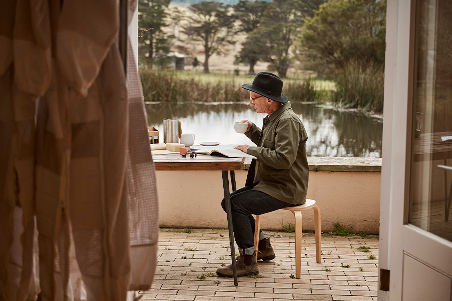Hamish seated at an outdoor table, drinking from a cup and reading, with a peaceful pond and trees visible in the background through an open doorway.