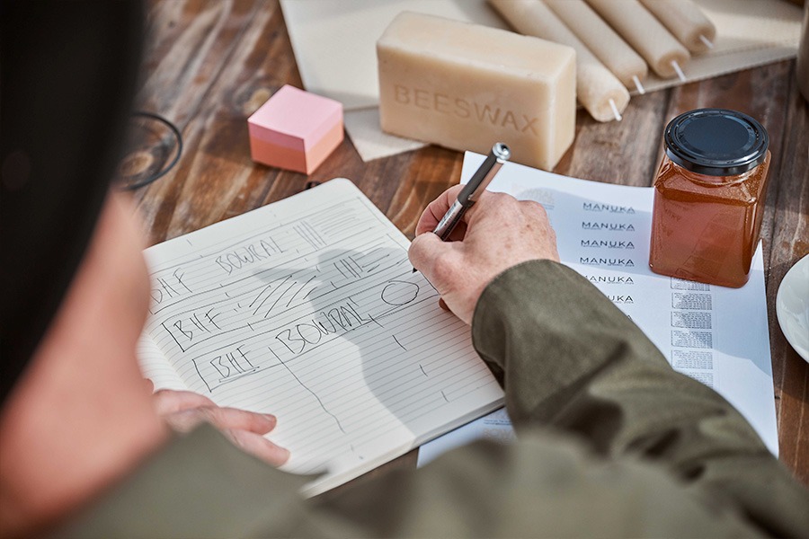 An over-the-shoulder view of Hamish writing in a notebook on a wooden table surrounded by beeswax, honey jars, candles and printed labels.